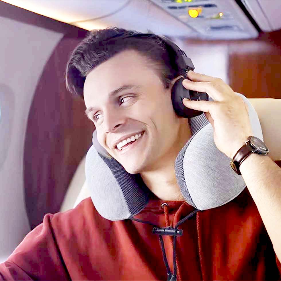 Hombre feliz viajando en avión, usando almohada ergonómica Ergo Travel Pillow y audífonos, expresando comodidad y disfrute durante el trayecto